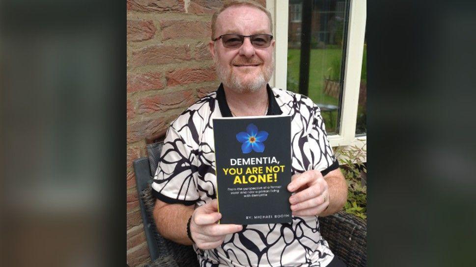 Mr Booth sitting on a chair in a black and white t-shirt and wearing glasses as he holds a copy of his book Dementia, You Are Not Alone.