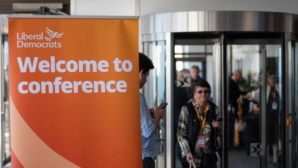 Lib Dem members arriving at the conference in Brighton, through a revolving door with a large orange Lib Dem banner to the left hand side, reading: "Welcome to conference".