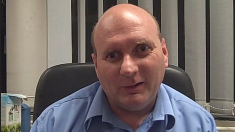 Jon Tandy is wearing a blue shirt and is sitting in a black office chair with a window and white blinds behind him.