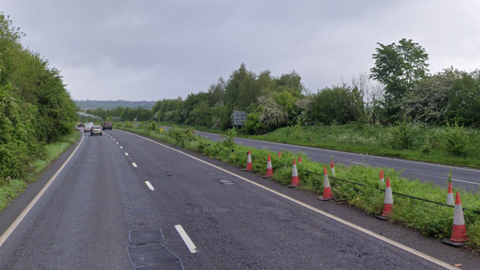 A dual carriageway with a few potholes visible and a line of orange cones in the central reservation. There are trees and bushes adjacent to the road.