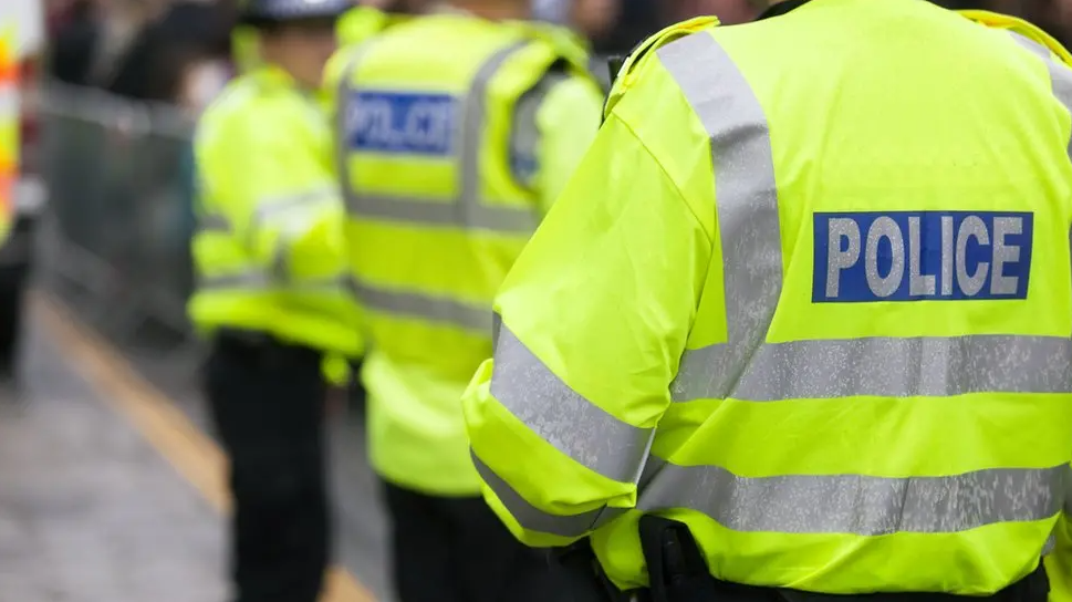 A photo taking from behind a police officer who is wearing a yellow hi-vis jacket with the word POLICE written on it. Their face, nor their head can be seen.