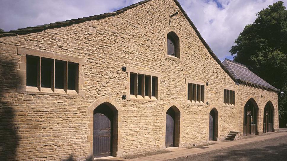 External view of the stone-built long barn at Gawthorpe Hall in Padiham. The stone is yellowy in colour and the building has four sets of four rectangular windows, three arched doorways and a further section at the end with larger arched doors.