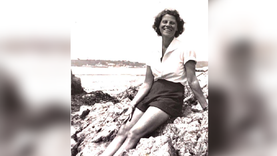 A black and white photo of a young woman in a white shirt and dark shorts sitting on some rocks. 