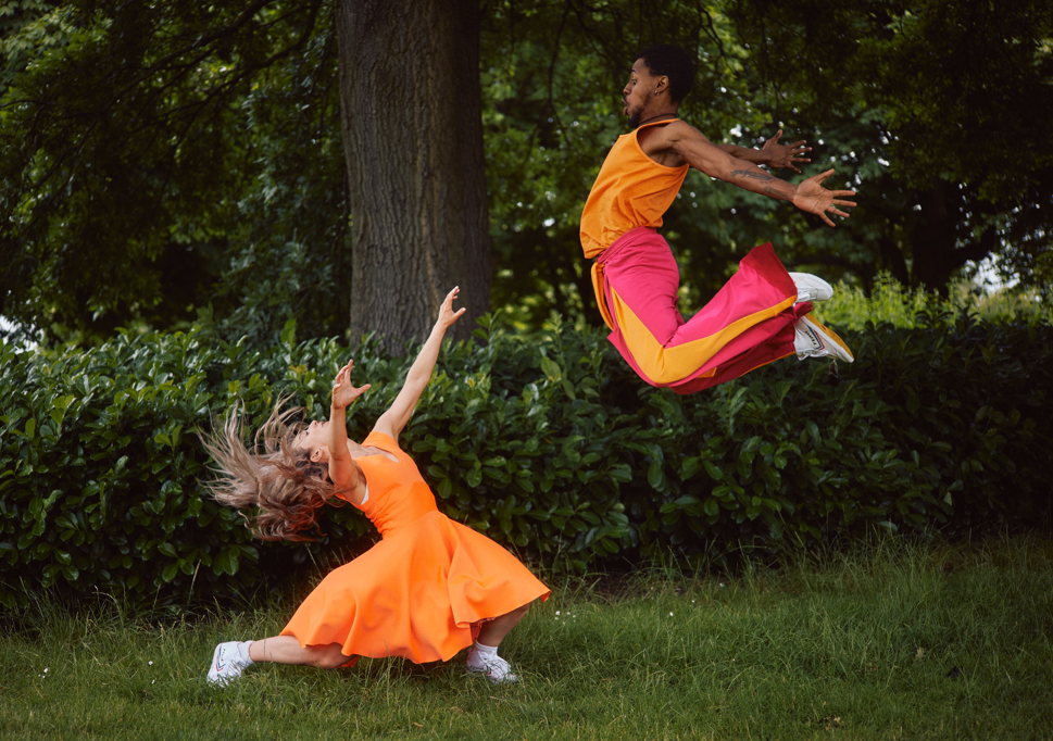 Two professional dancers are wearing orange and pink costumes. One has jumped in the air and the other is flipping her hair.