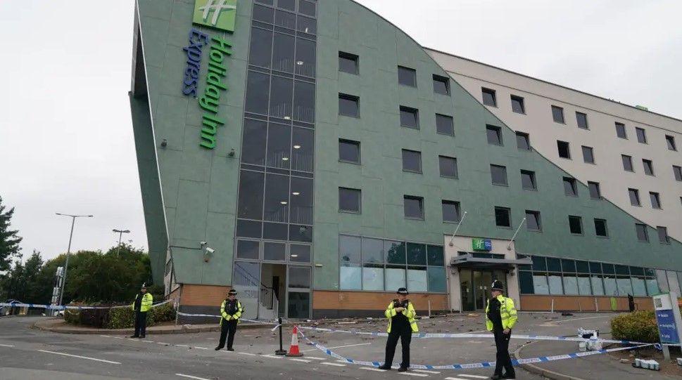 Four police officers standing next to a cordon next to the Holiday Inn Express in Tamworth. The building is green and beige and has the logo on the left. Debris can be seen next to the hotel, behind the cordon.
