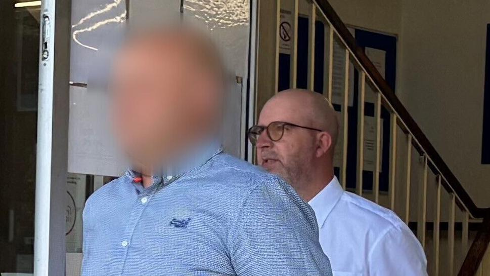 Andrew Wickenden wearing glasses and a white shirt leaving court behind another man who is wearing a blue shirt. The face of the second man, in the foreground, is blurred out.