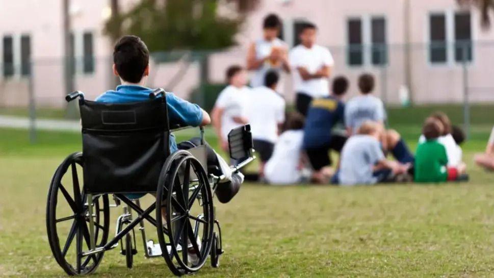 A boy sat in a black wheelchair in a field, looking at a group of young people sat and stood together in the distance