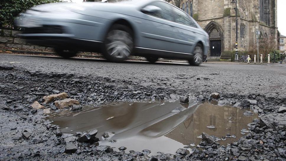 A blurred silver/blue car passing a pothole which is in the foreground full of water and loose stones. There is a church in the background