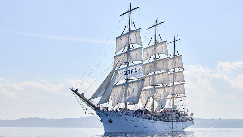 The tall ship, which is white and has three masts at full sail.