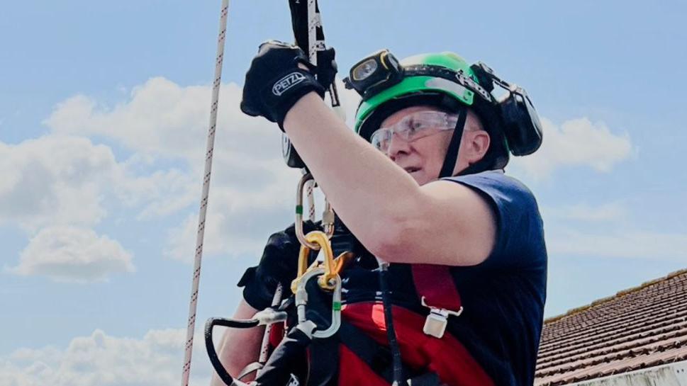 Andrew Gibson abseiling. He is wearing a blue T-shirt, a safety helmet and gloves