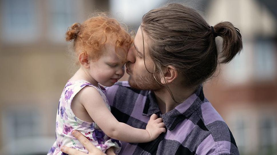 James, who's wearing a dark and light purple checked shirt, kissing his young daughter Violet, who has red hair and is wearing a purple and white floral dress.