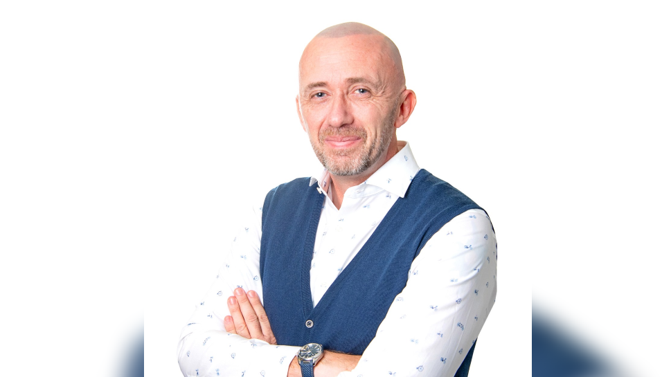 A man with his arms crossed, wearing a white shirt and a blue sleevless cardigan smiles at the camera. He has a shaved head and a grey beard