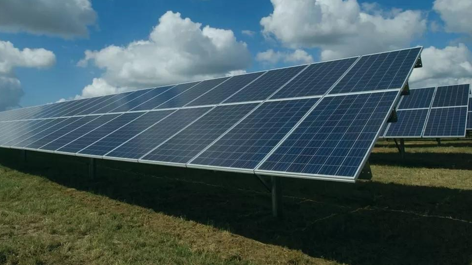 A large solar panel in a green field. The panel is facing the sky which has a number of clouds in it. 