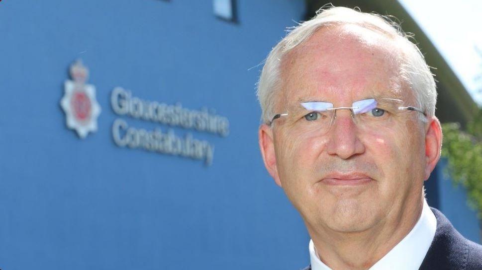 Chris Nelson standing in front of a blue building which says 'Gloucestershire Constabulary' on it. He has white hair, is clean shaven and wearing clear framed glasses.