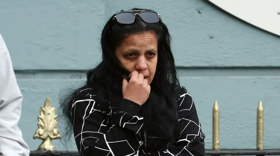 A woman, with long black hair, wearing a black top with white lines on it. She is holding one hand up to her mouth. There is a blue wall in the background.