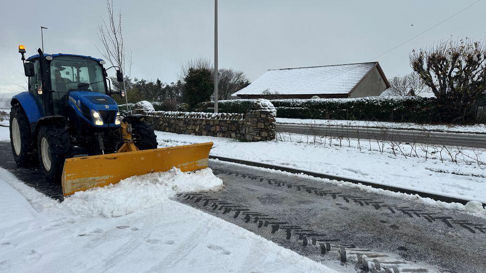 A snow plough is photographed on a snow-covered road, its yellow plough moving snow from the road.