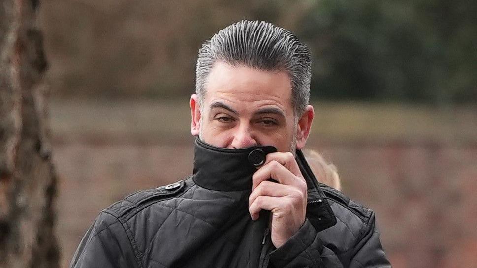 Christopher Nunn wearing a black coat. He is pulling the collar up to hide part of his face. He has short, slicked-back black and grey hair.