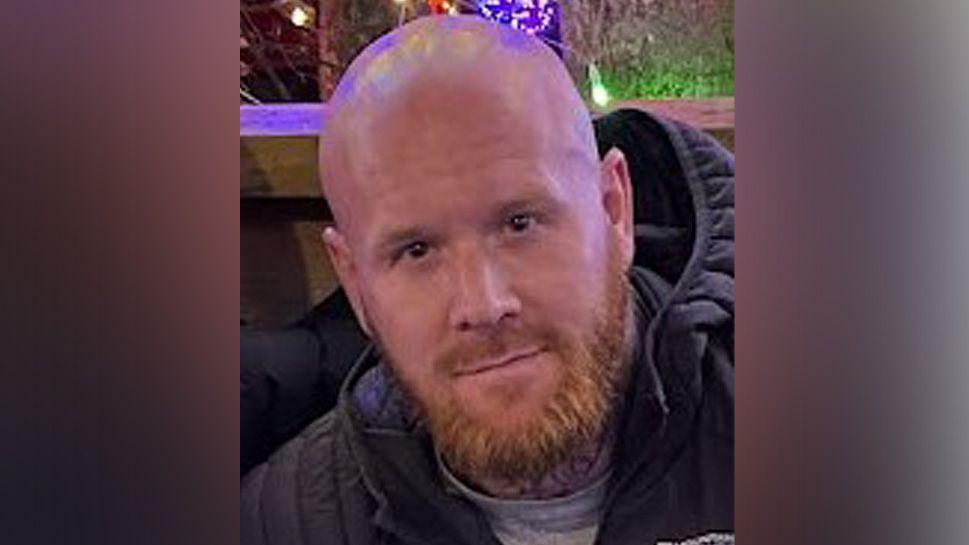 Lenny Scott, who has brown eyes, shaven head and ginger beard and is wearing a navy blue puffer coat and grey t-shirt. 