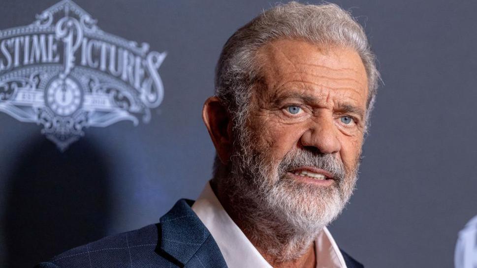 Mel Gibson, wearing a dark blazer and white shirt with an open collar, with grey hair and a grey beard, poses for photographs at a screening in Los Angeles
