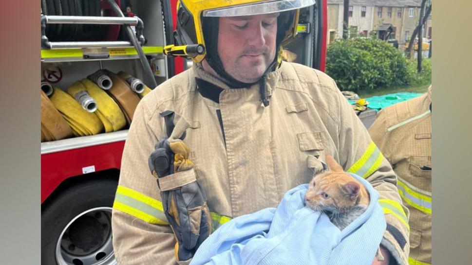 A firefighter in uniform holds the ginger cat which is wrapped in a blue blanket