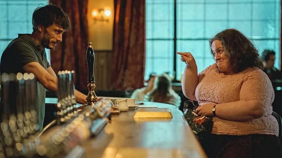 Richard Gadd plays barman Donny and Jessica Gunning his stalker Martha in the TV series Baby Reindeer. Image shows a man with dark hair leaning over a bar and a woman sat at the bar, smiling and pointing at him.