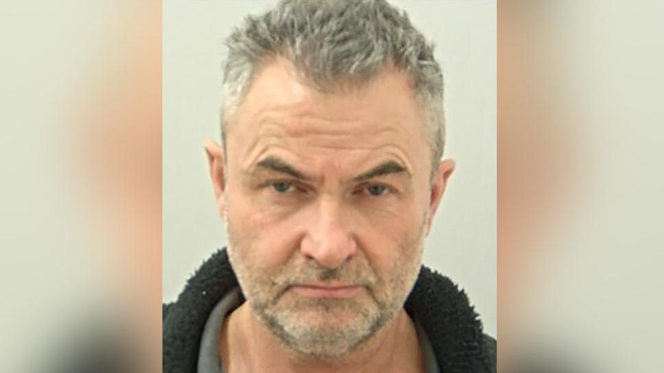 Police custody image of Robert Easom with grey hair and beard wearing a grey top and black fleece.