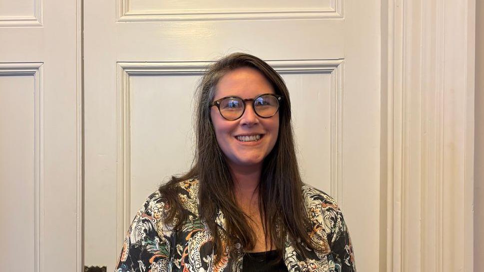 Charlotte Traynor, a woman with long dark hair, she wears large round glasses and has a bright smile, she wears a patterned white and black jacket.