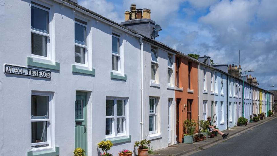 A row of colourful houses on Athol Terrace in Castletown. The houses' front doors open onto the pavement, which runs alongside a road. Many have boxes containing flowers or small bushes either side of the doorway and a person in shorts is sitting outside one of the houses halfway along the terrace.