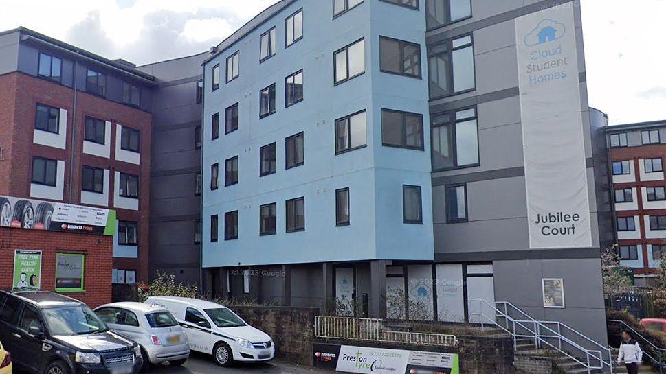 Street view image of Jubilee Court, a four-storey building made of three blocks, a red brick one to the left, a pale blue one in the middle and a grey block to the right. There is a small car park to the front with three vehicles