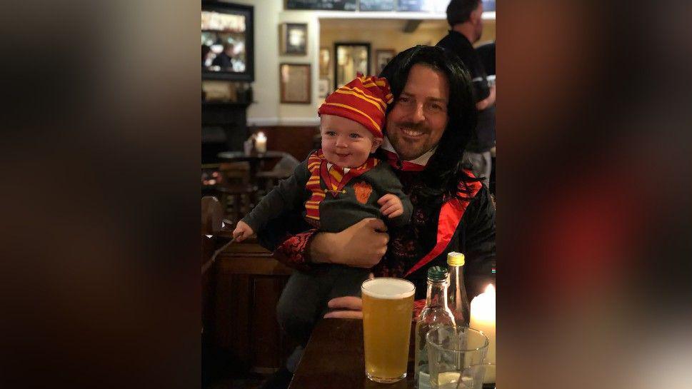 Matthew holding Richard as a toddler in a pub. Matthew is wearing a black fancy dress outfit and Richard is dressed up as a mini Harry Potter in a school uniform, wearing a red and yellow hopped beanie hat and holding a wand.