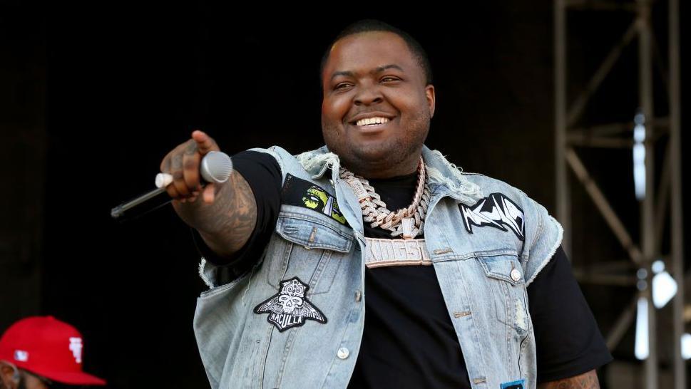 Rapper Sean Kingston is seen on stage holding a microphone and pointing out into the audience. He is smiling and wearing a black T-shirt with a jean vest over it. 