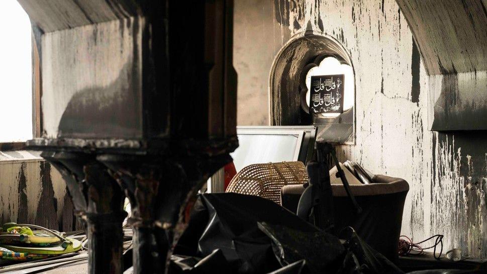 A photograph taken from inside a mosque. The walls are damaged and blackened by smoke. There is furniture in disarray. On the wall is an Islamic motif.
