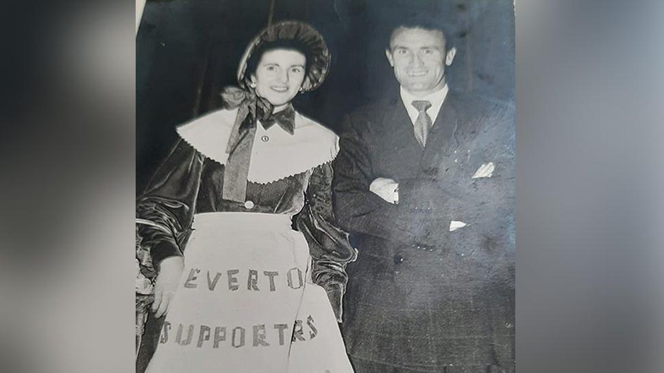 Everton FC toffee lady Mary Morgan (nee Gorry) wearing Victorian style confectionary attire with white pinny and bonnet with Tommy Eglington who played for Everton wearing a suit taken between 1953 and 1956.