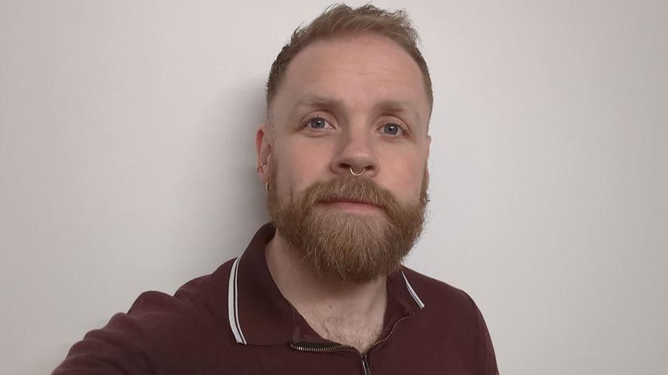 Liam Behan smiling at the camera. He is wearing a purple shirt and has fair hair, a beard and a nose ring.