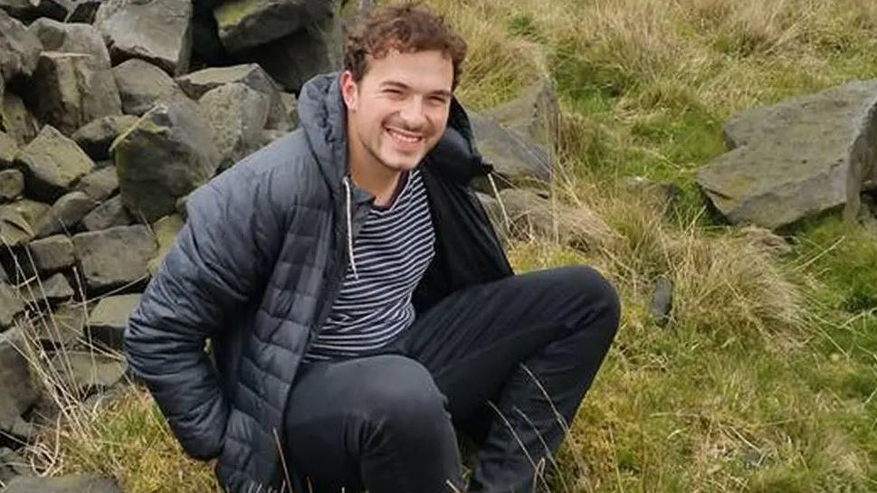 Jack Ritchie, a man in his 20s with dark hair and wearing black jeans, a striped T-shirt and a black jacket, sitting down in a field next to a stone wall.