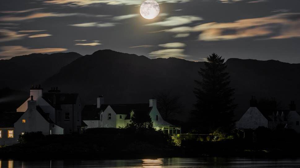 Full moon shines over white cottages and mountains, reflecting on a still lake.