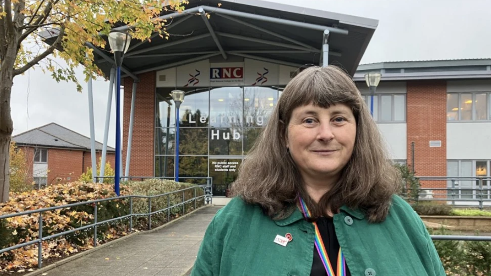 A woman with shoulder-length brown-grey hair and wearing a green jacket stands in front of a glass-fronted building labelled "RNC Learning Hub"