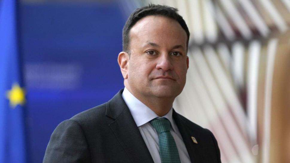 A man with short black hair stares into the camera. He has a lightly checked dark grey suit, and a grey tie. Behind him, out of focus, the blurry outline of a European flag can be seen.