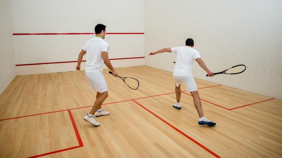 Two men playing squash in a sports hall, wearing white shorts and T-shirts and trainers.