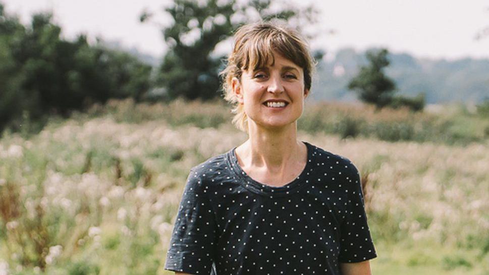 Daisy Greenwell smiles at the camera while standing in a field. She has brown hair which has been tied up and wears a polka dot top.