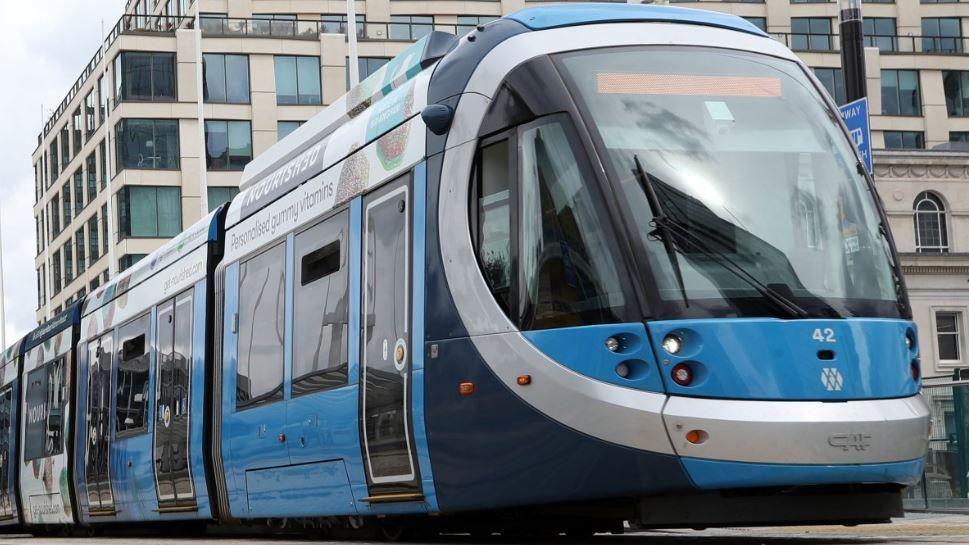 A tram in Birmingham city centre. It is blue and there is a large building behind it. 