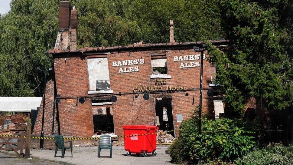 The remains of a pub building after a fire. The brick facade of the building is still intact but the roof is totally destroyed, and the inside of the building seen through windows and doorways is completely empty. The building has subsidence and very prominently leans to the left. On the front of the building is a sign that says The Crooked House, as well as a sign for Banks's Ales.

