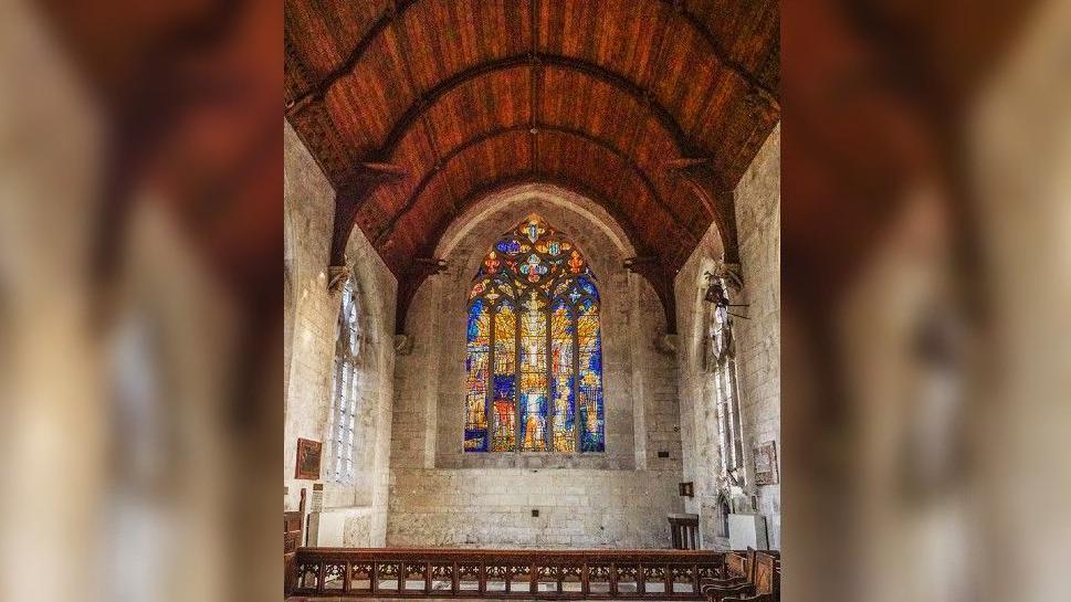 A view of the whole east window in an otherwise plain chapel with unadorned stone walls and a wooden roof