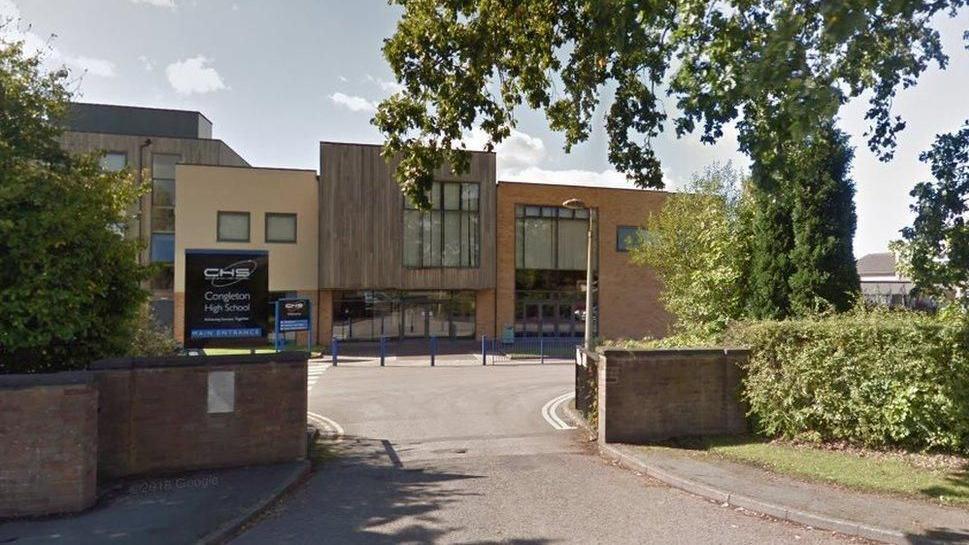 The front of a school building with a driveway leading up to it. There are blue bollards directly infront of a brown reception area