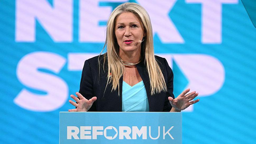 Laura Anne Jones wearing a black jacket and blue top, standing at a podium with her hands raised. She has a white and blue backdrop behind her and is wearing a microphone.