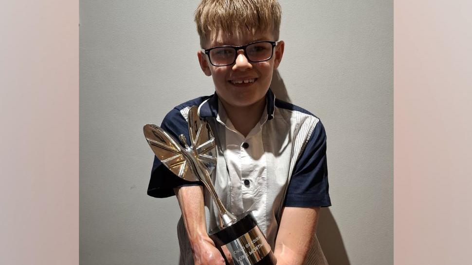 Luke Mortimer is pictured against a grey background wearing a black and white shirt and holding his large Pride of Britain award in the shape of a silver winged woman.