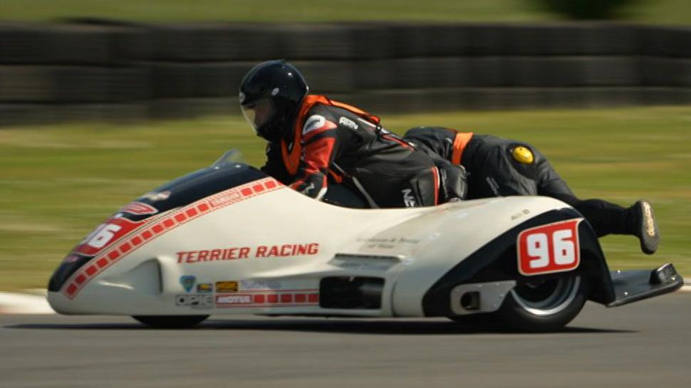 A white sidecar, with red writing reading 'Terrier Racing' on the side, and the number 96 printed above the wheel arch. The driver is wearing black and red leathers, a black helmet, and an orange hi-vis vest. To the right, you can see the passenger leaning over the right hand side of the sidecar to counterbalance it. She's also wearing black leathers, a black helmet, and an orange hi-vis vest. The sidecar and the racers are in focus, but the background is blurred.