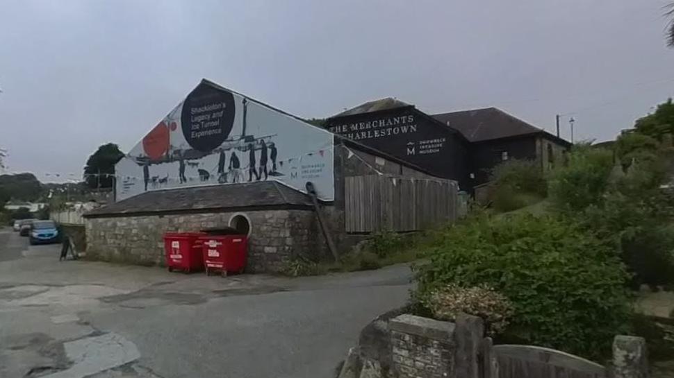 Google Street View image of the Shipwreck Treasure Museum in Charlestown. A mural about an Ernest Shackleton exhibition is on a gable wall. Two large red bins are just below the mural.