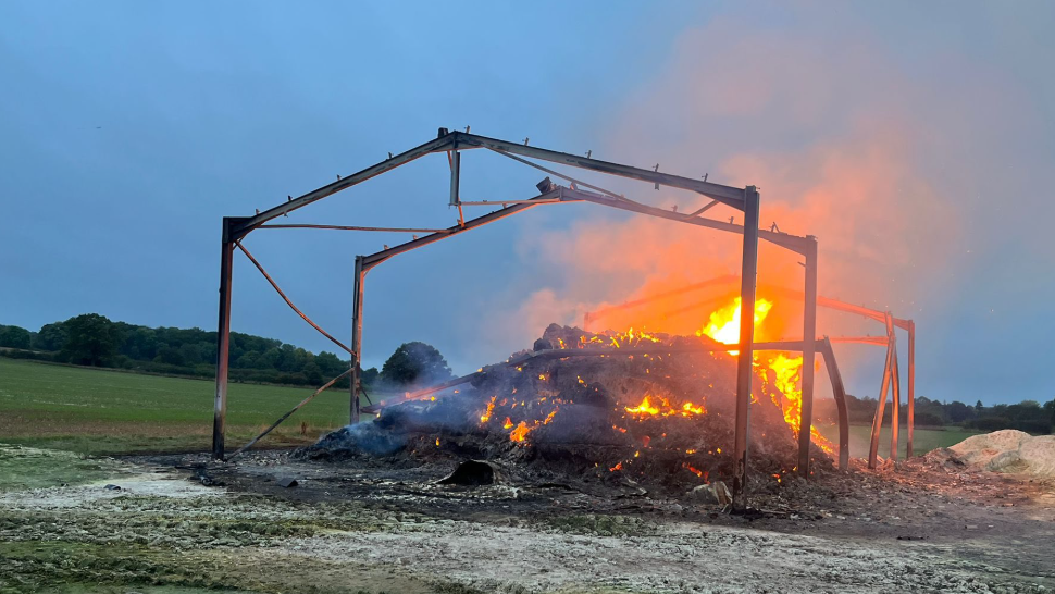 The remains of a large metal building, with large flames rising from burnt material inside it. There is a field behind the building, and burnt ground in front.