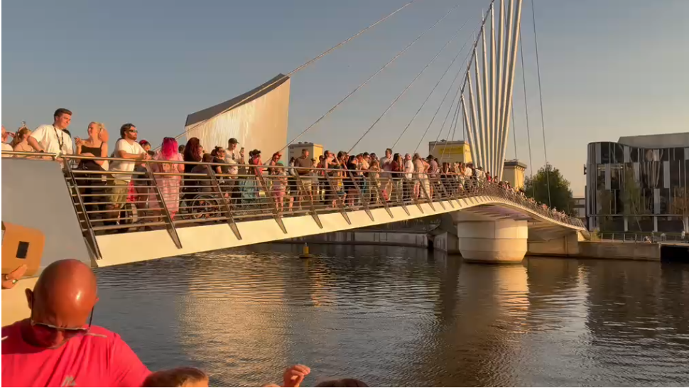 Image shows over a hundred people standing on a bridge at MediaCity, looking up into the sky